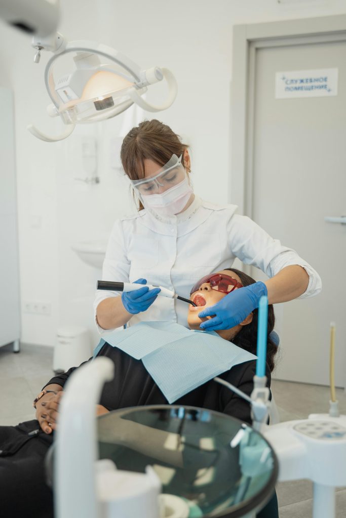 A dentist uses a dental tool on a patient's mouth in a modern dental office, showcasing proper safety measures with masks and protective gear.